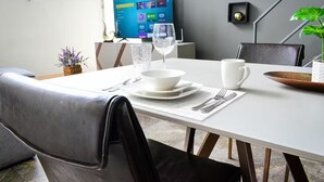 Ein moderner Essbereich mit einem weißen Tisch, gedeckt für zwei Personen, einem Glas Wasser, einem Weinglas und einer Schüssel. An der Wand ist ein Flachbildfernseher angebracht, der eine Spieloberfläche zeigt.