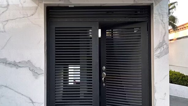 Ein moderner Gebäudeeingang mit einem schwarzen Metallgitter, einem kleinen Fenster und einem Betonboden.