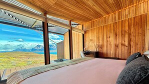 A bedroom with a large bed, wooden walls, a desk, and a view of mountains and a lake.