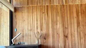 A modern interior with a wooden slatted ceiling, a dark wooden bench, a desk with a vase of dried plants, and a grey upholstered chair.