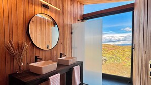 A modern bathroom with wooden walls, a large mirror, two sinks, and a view of a mountainous landscape through an open door.
