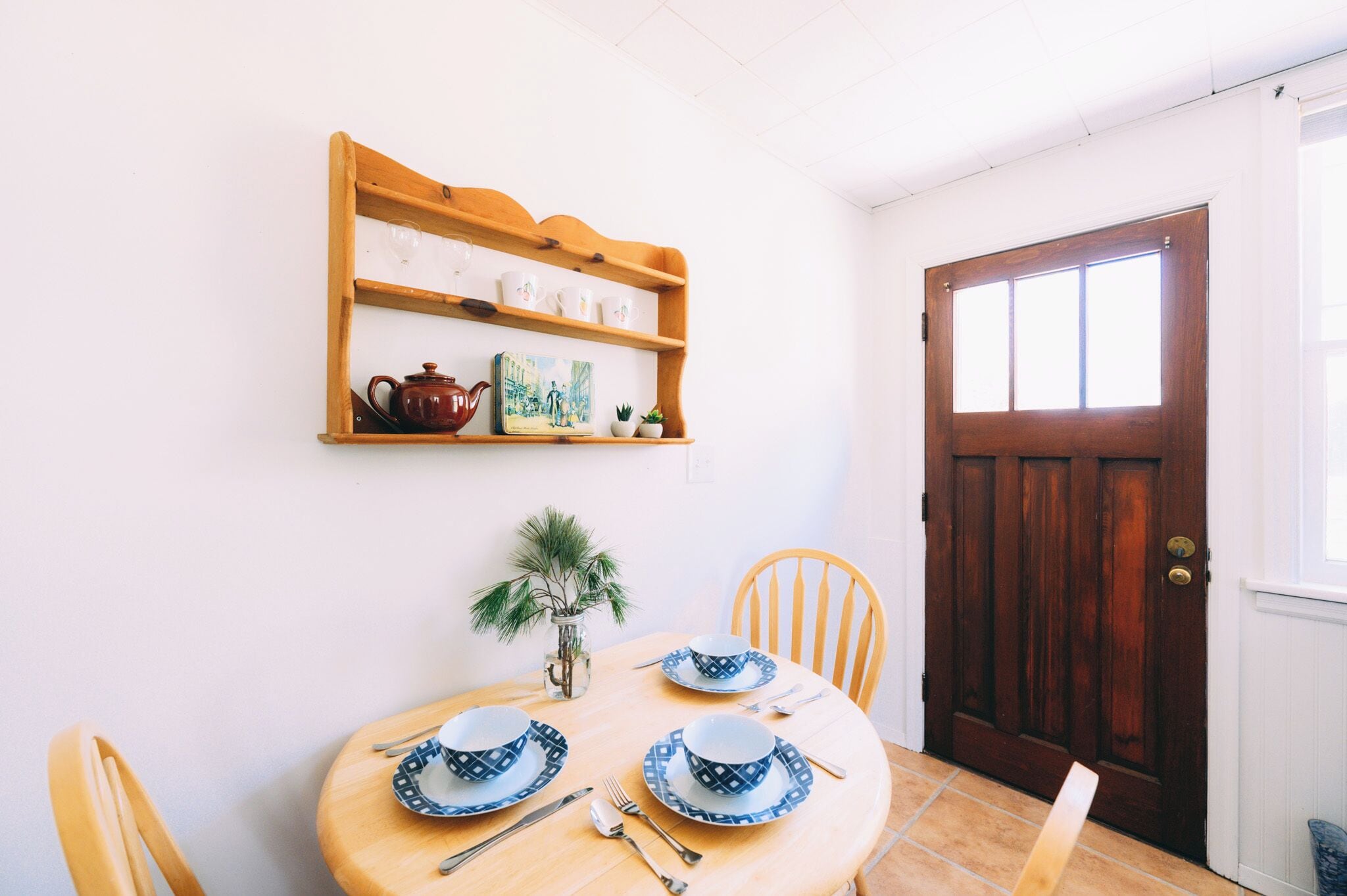 Un coin repas avec une table en bois dressée pour quatre personnes, une étagère en bois au-dessus, ornée d’objets décoratifs, et une porte en bois sur la droite.