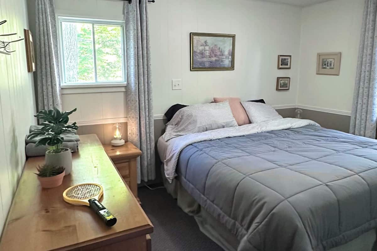 A bedroom with a bed, a dresser, a window with curtains, a ceiling fan, and a wall-mounted picture.