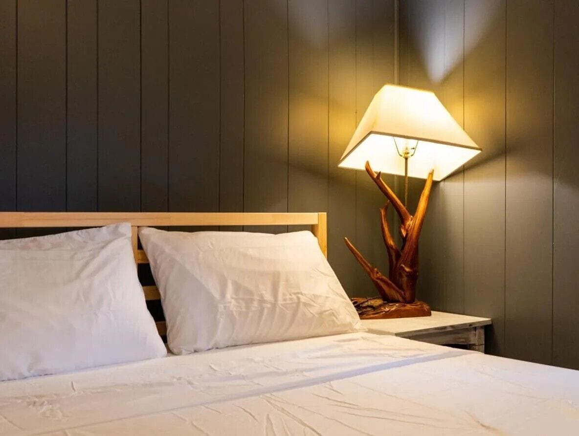 Una cama bien tendida con sábanas blancas y dos almohadas, un cabecero de madera y una mesita de noche con una lámpara de base de madera y pantalla blanca.