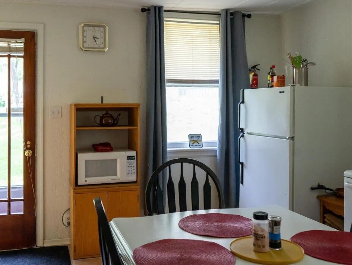 A small kitchen with a microwave, a refrigerator, a wooden shelf, and a window with curtains.