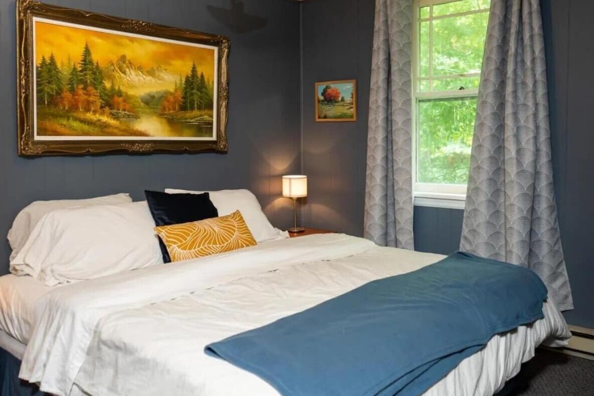 A bedroom with a bed, a painting, a lamp, and a window with curtains.