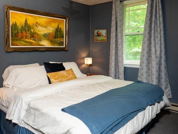 A bedroom with a bed, a painting, a lamp, and a window with curtains. - Falcon Retreats (Sharbot Lake)