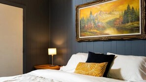 Un lit bien fait, avec une literie blanche et bleue, une table de chevet avec une lampe, et un tableau encadré représentant une scène de forêt accroché au mur.