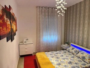A bedroom with a bed, a nightstand, a dresser, and a window with curtains. - Casa Gabriela: A Peaceful 2BR Gateway | Private Parking, AC, Near Beach (Viddalba)