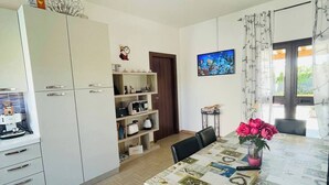 Une cuisine moderne avec des meubles blancs, une table à manger recouverte d’une nappe à motifs et un téléviseur fixé au mur.