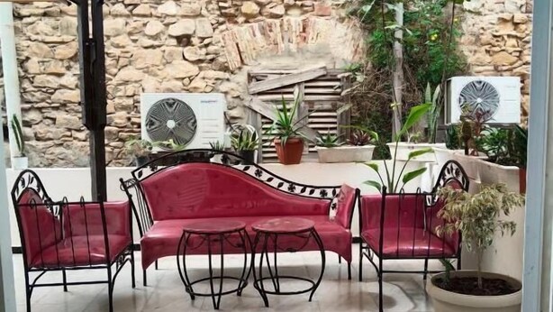 Un'area patio esterna con un muro in pietra sullo sfondo, un divano rosso, due tavoli rotondi e un ombrellone color beige.