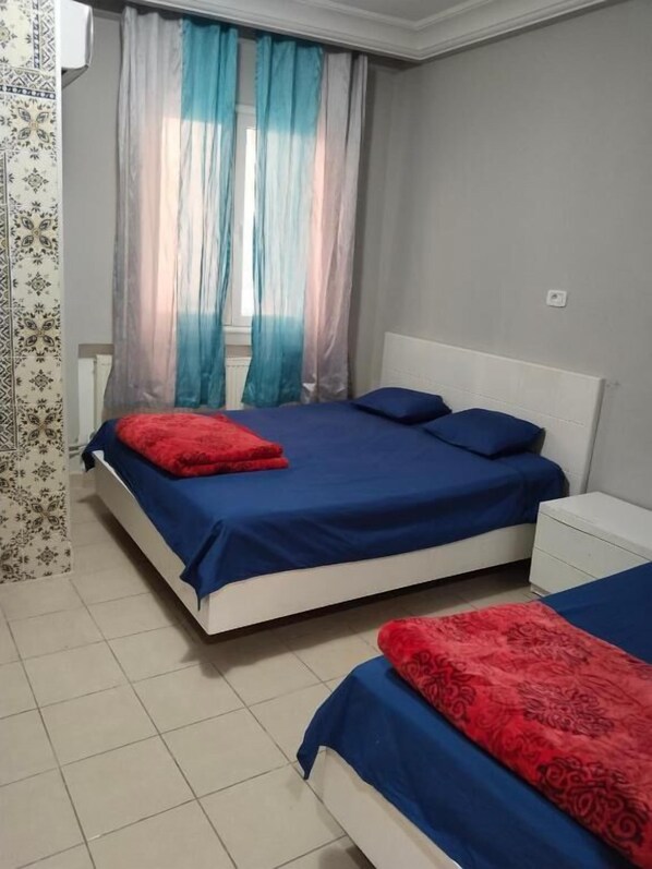Ein Schlafzimmer mit zwei Betten, beide mit blauer Bettwäsche und roten Kissen. Links befindet sich ein Fenster mit durchsichtigen Vorhängen und einem gemusterten Vorhang.