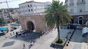 Una piazza pubblica con un arco imponente, palme e una varietà di pedoni e auto parcheggiate.