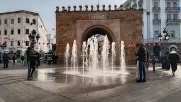 A public square with a central fountain, a stone archway, and surrounding buildings.