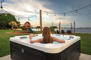 Eine Person entspannt sich in einer Whirlpool-Badewanne mit Blick auf einen See und einen Spielplatz im Hintergrund.