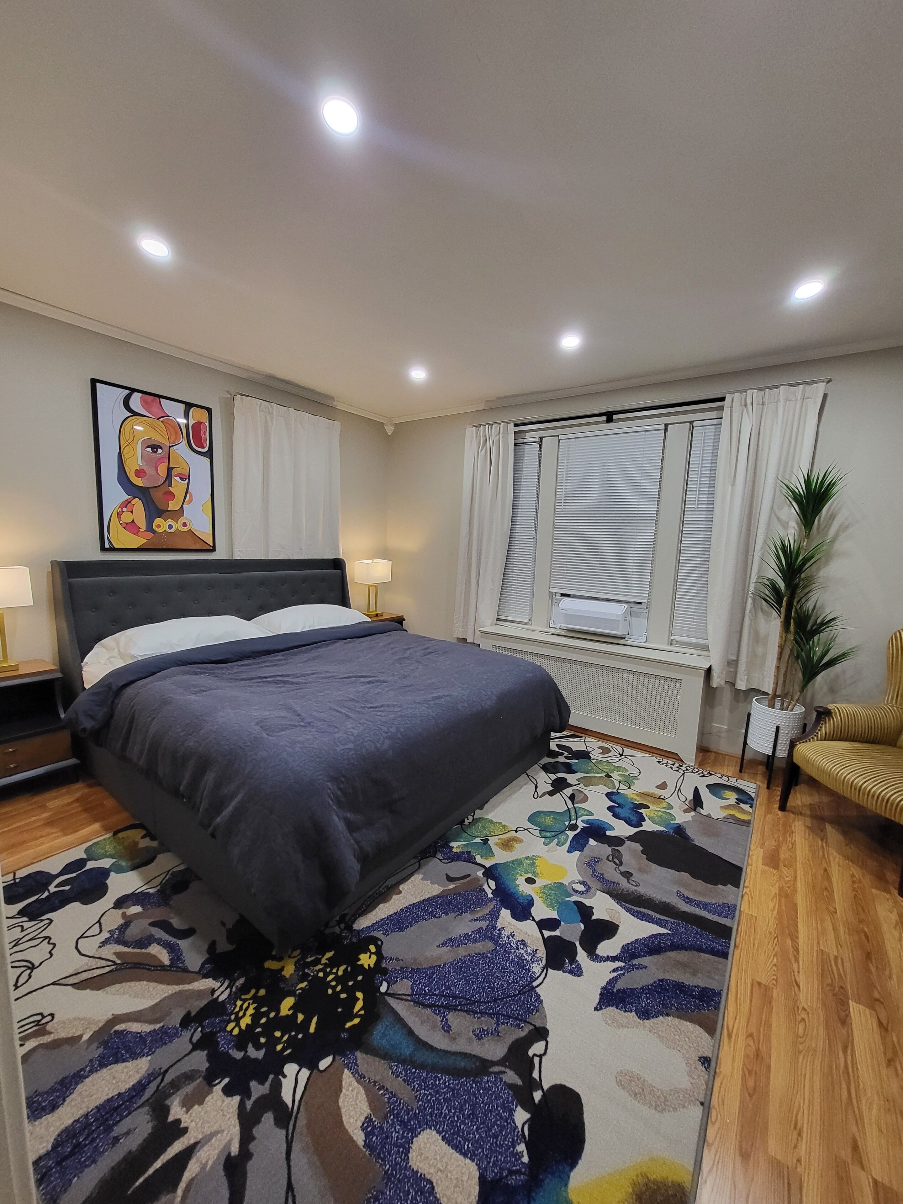 Een slaapkamer met een bed, nachtkastje, lamp, raam met zonwering, radiator, kamerplant en een stoel.