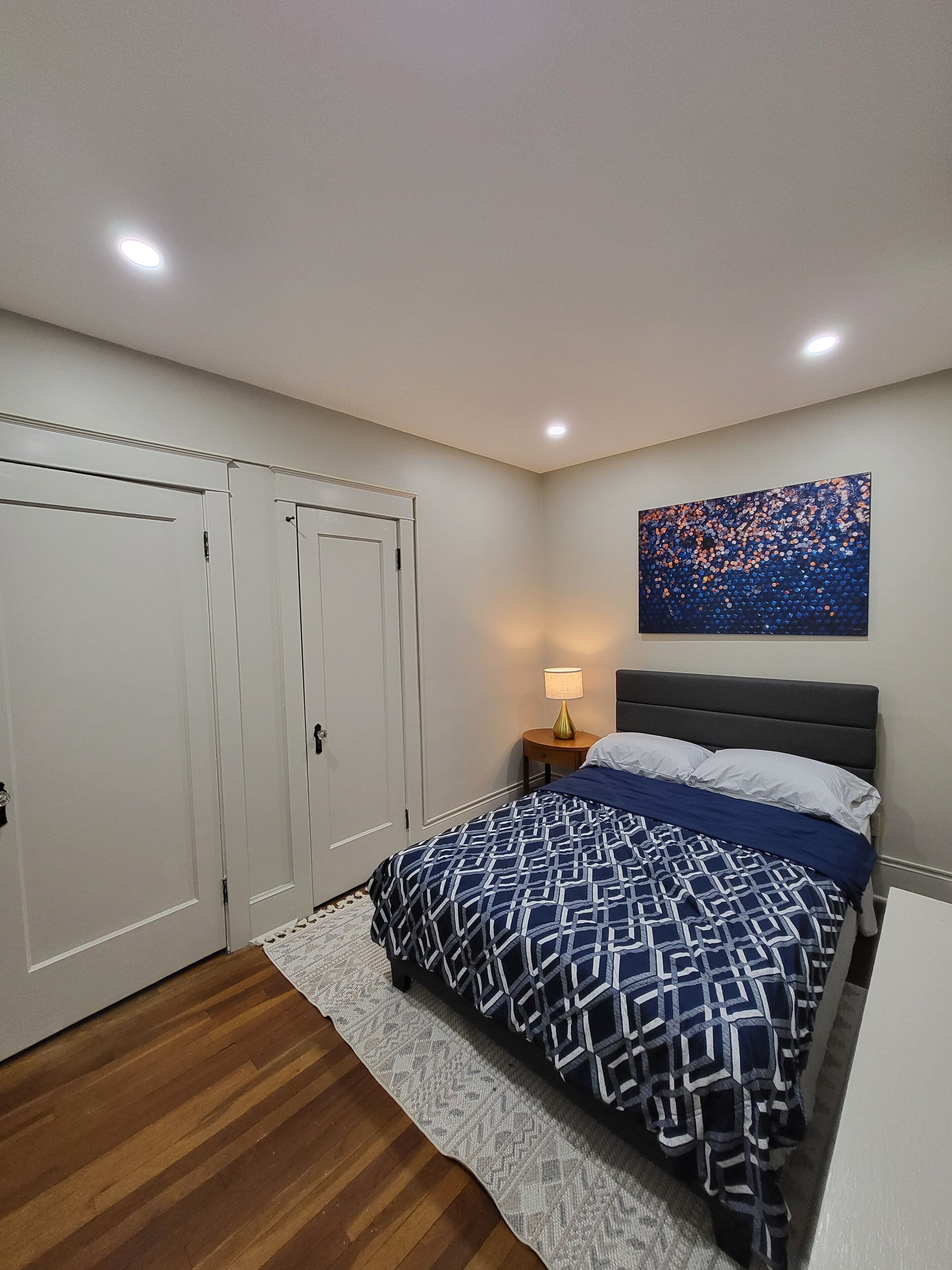 A bedroom with a bed, a nightstand, a lamp, a painting, and a wardrobe.