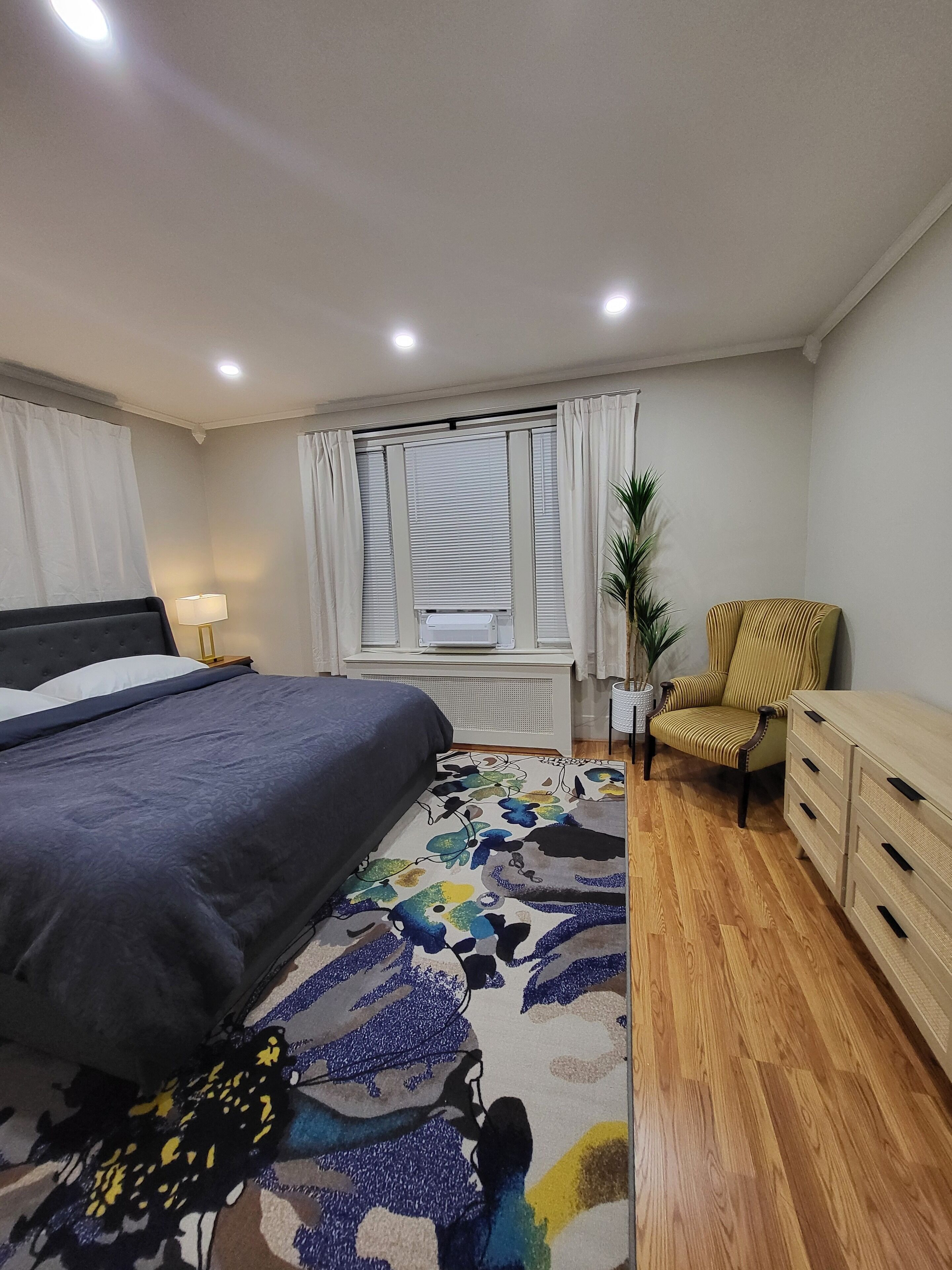 A bedroom with a bed, a chair, a dresser, a radiator, and a window with curtains.
