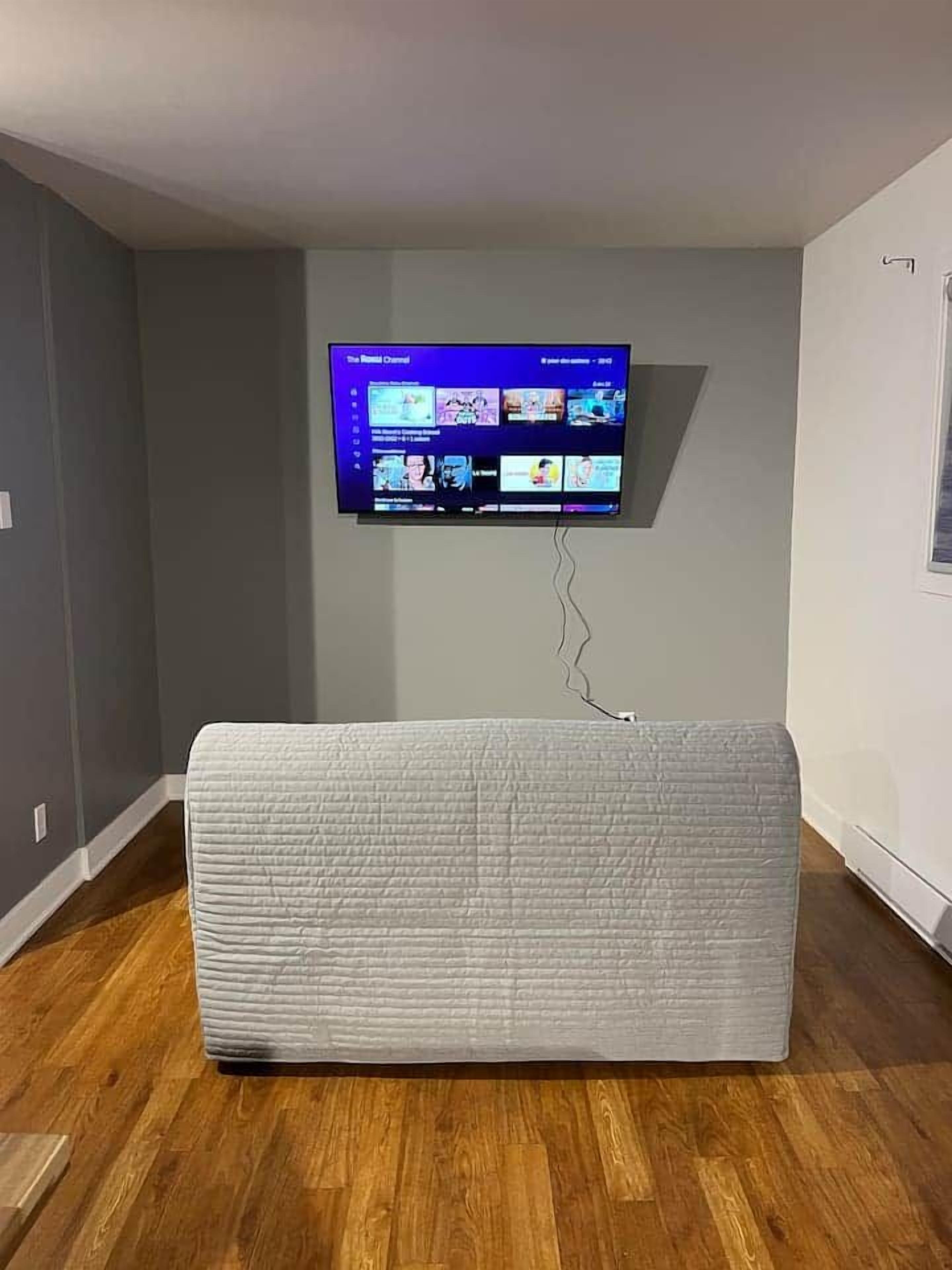 A room with a flat-screen TV mounted on the wall, a white sofa, and wooden flooring.