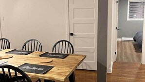 Un coin repas avec une table en bois et des chaises noires, une porte blanche et un couloir menant à une autre pièce.