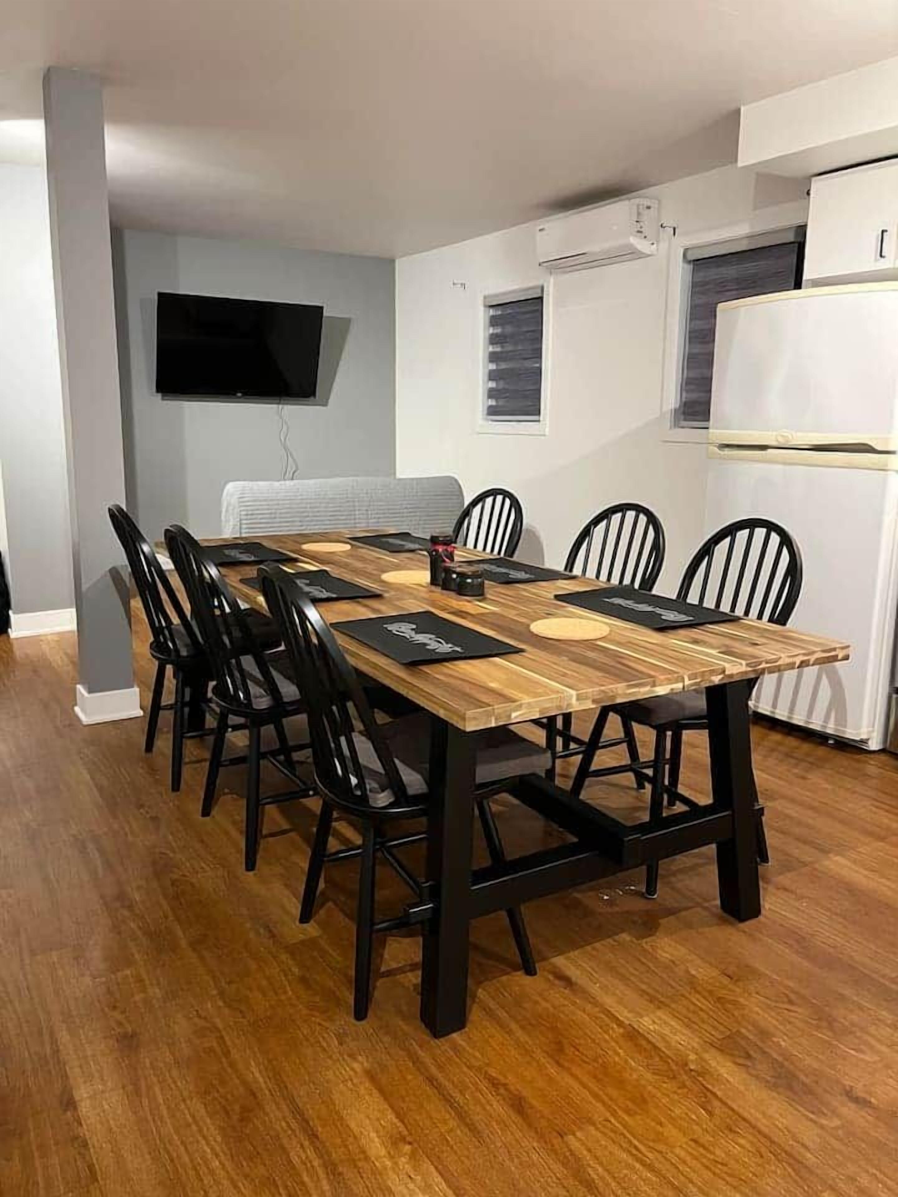 A dining area with a long wooden table, black chairs, a flat-screen TV mounted on the wall, and a white refrigerator.