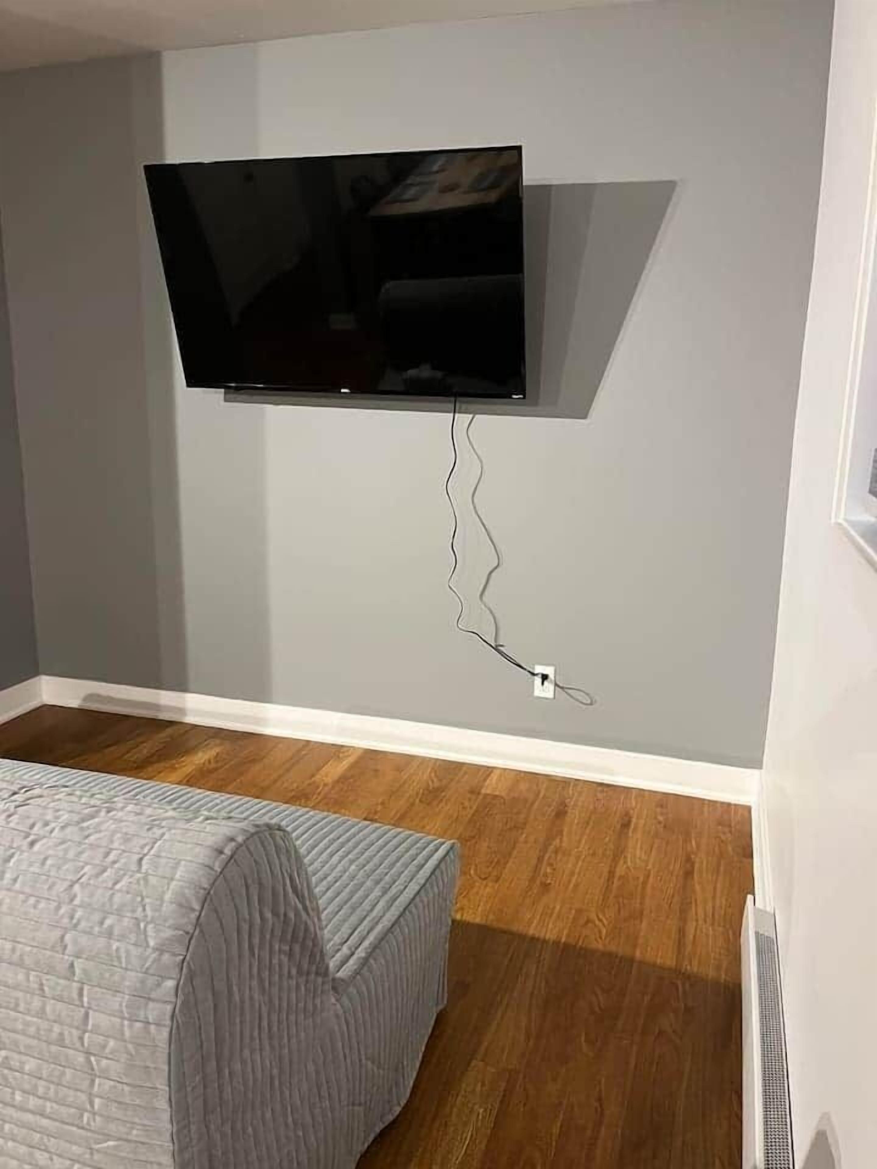 A room with a mounted TV, a bed with a grey quilt, wooden flooring, and a white wall.