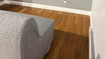 A room with a mounted TV, a bed with a grey quilt, wooden flooring, and a white wall.