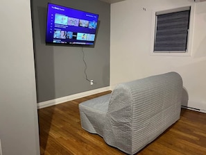 Habitación con una televisión de pantalla plana en la pared, un sofá tapizado en gris, suelo de madera y una ventana con persianas cerradas.