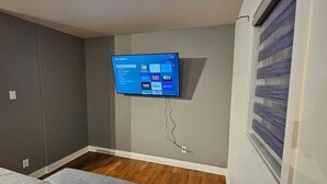 Une chambre équipée d’un téléviseur fixé au mur, qui permet d’accéder à des services de diffusion en continu, d’un lit et d’une fenêtre avec des stores.