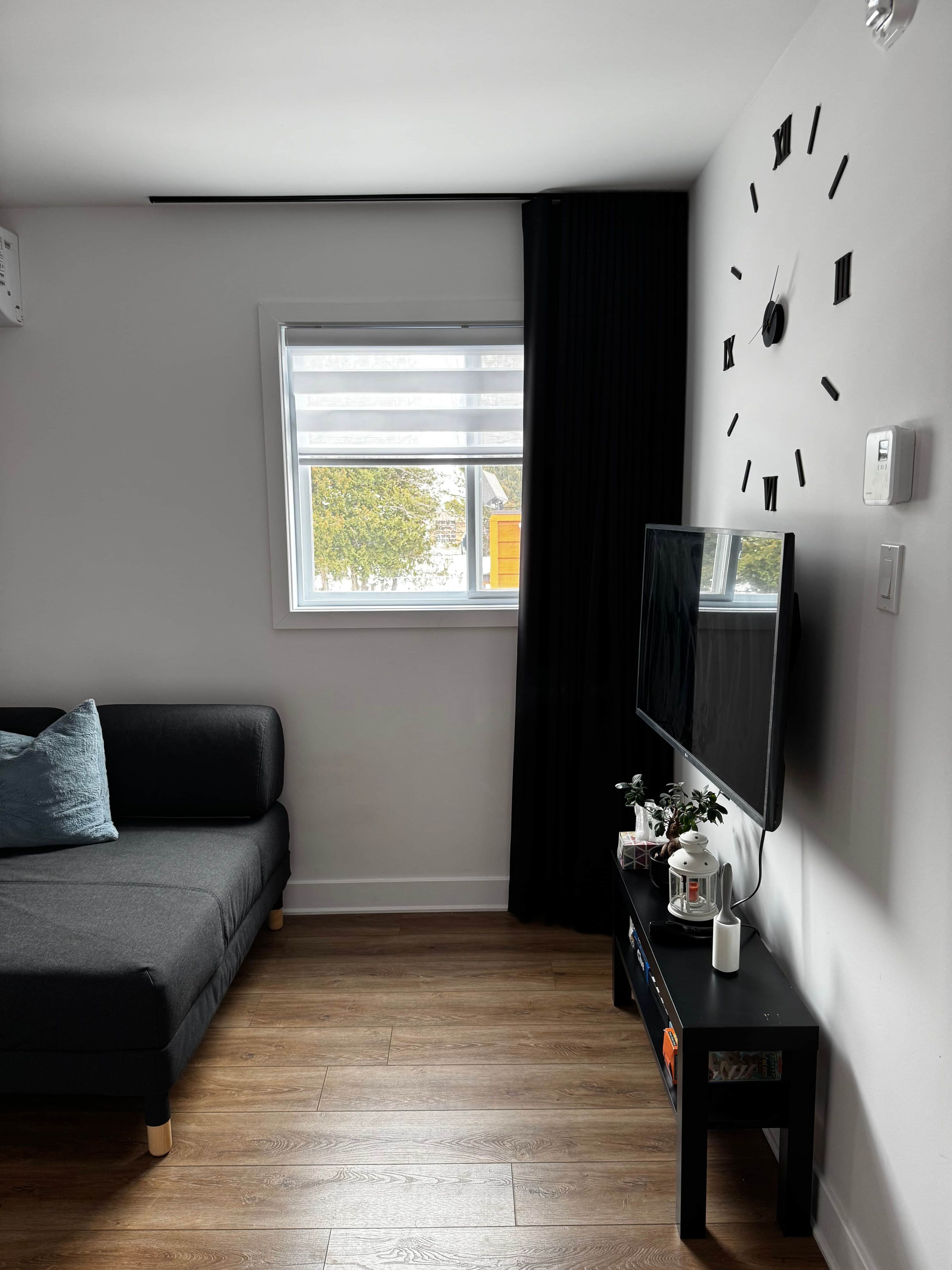 A modern living room with a black sofa, a flat-screen TV, a wooden floor, a window with blinds, and a wall-mounted clock.