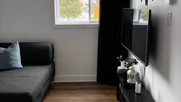 A modern living room with a black sofa, a flat-screen TV, a wooden floor, a window with blinds, and a wall-mounted clock.