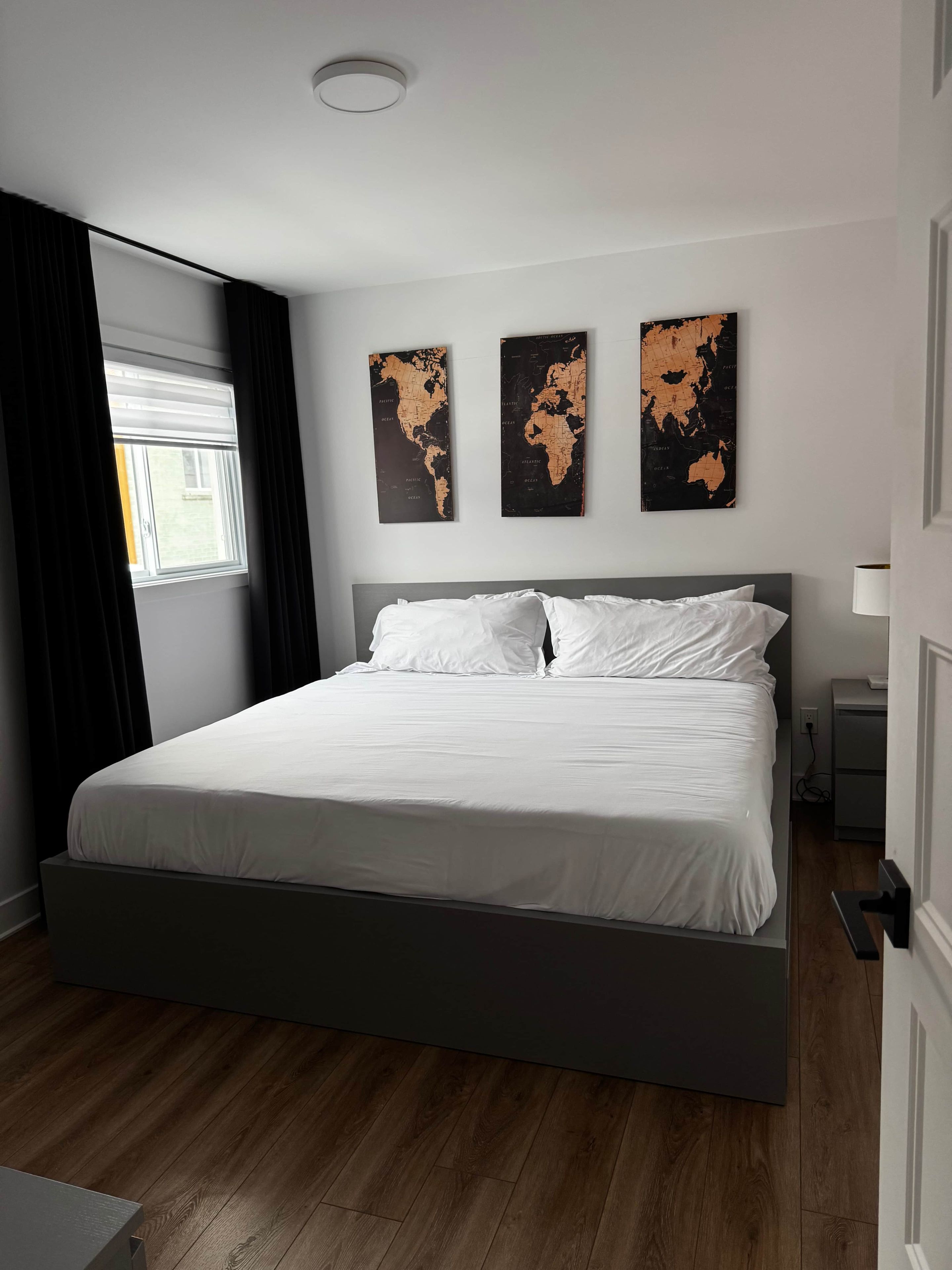 Une chambre moderne avec un grand lit, une fenêtre avec des rideaux et trois œuvres d’art représentant une carte du monde accrochées au mur.