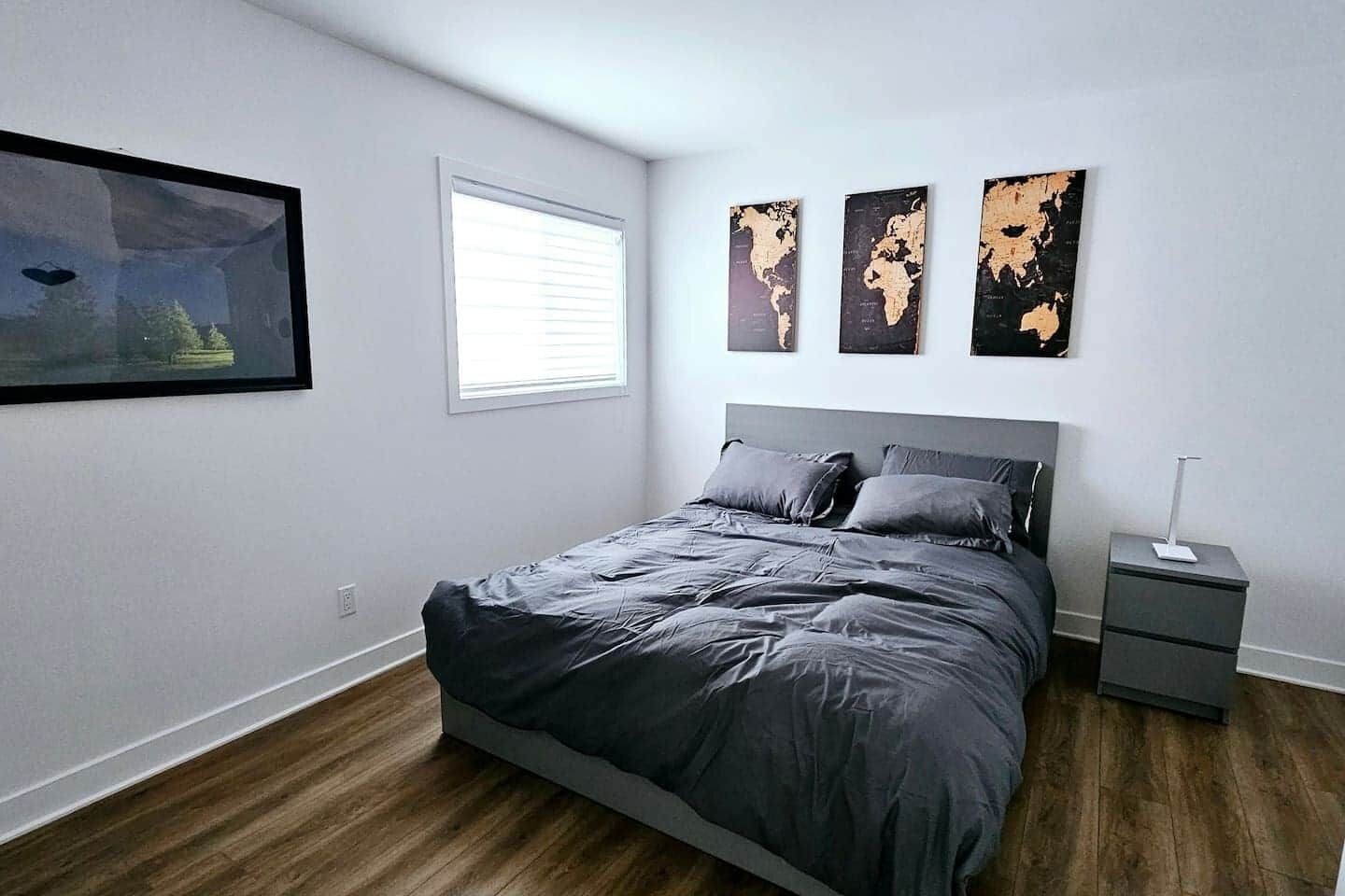 Een slaapkamer met een bed, nachtkastje, raam en drie wereldkaarten aan de muur.