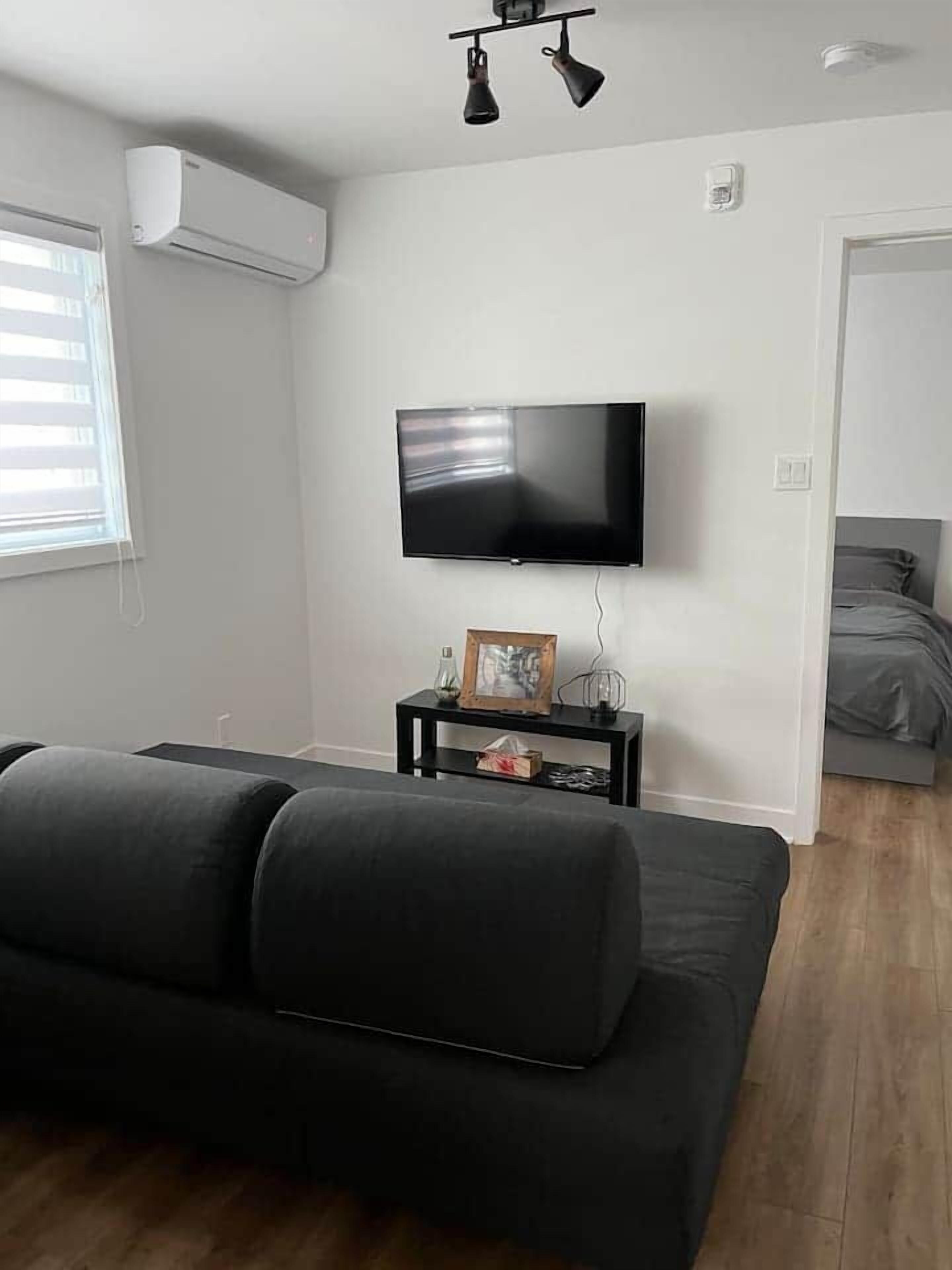 Een moderne woonkamer met een zwarte bank, een wandgemonteerde flatscreen-tv en een bijzettafeltje met een fotolijstje en decoratieve objecten.
