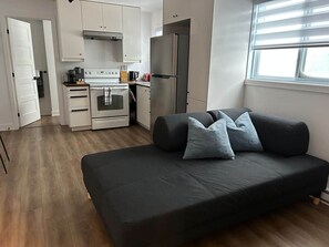 Un espacio habitable compacto con kitchenette, un sofá con cojines y un ventanal que proporciona luz natural.