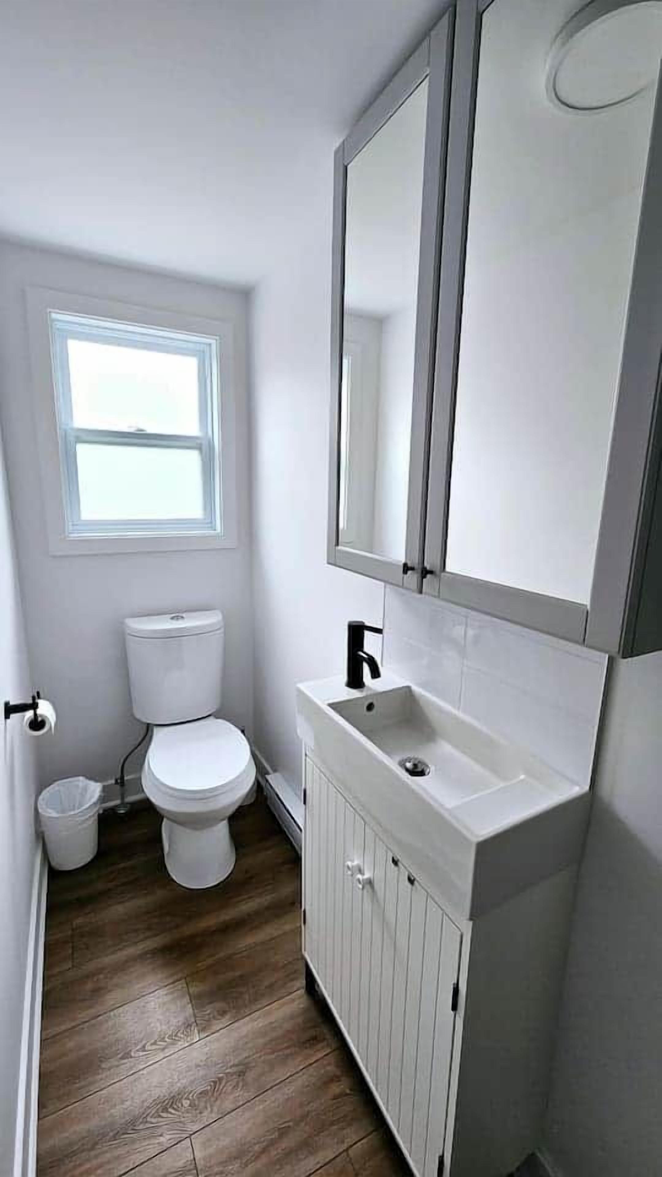 Et lille, hvidt badeværelse med toilet, vask og et spejl over vasken. Der er et vindue over toilettet, og gulvet er træ.
