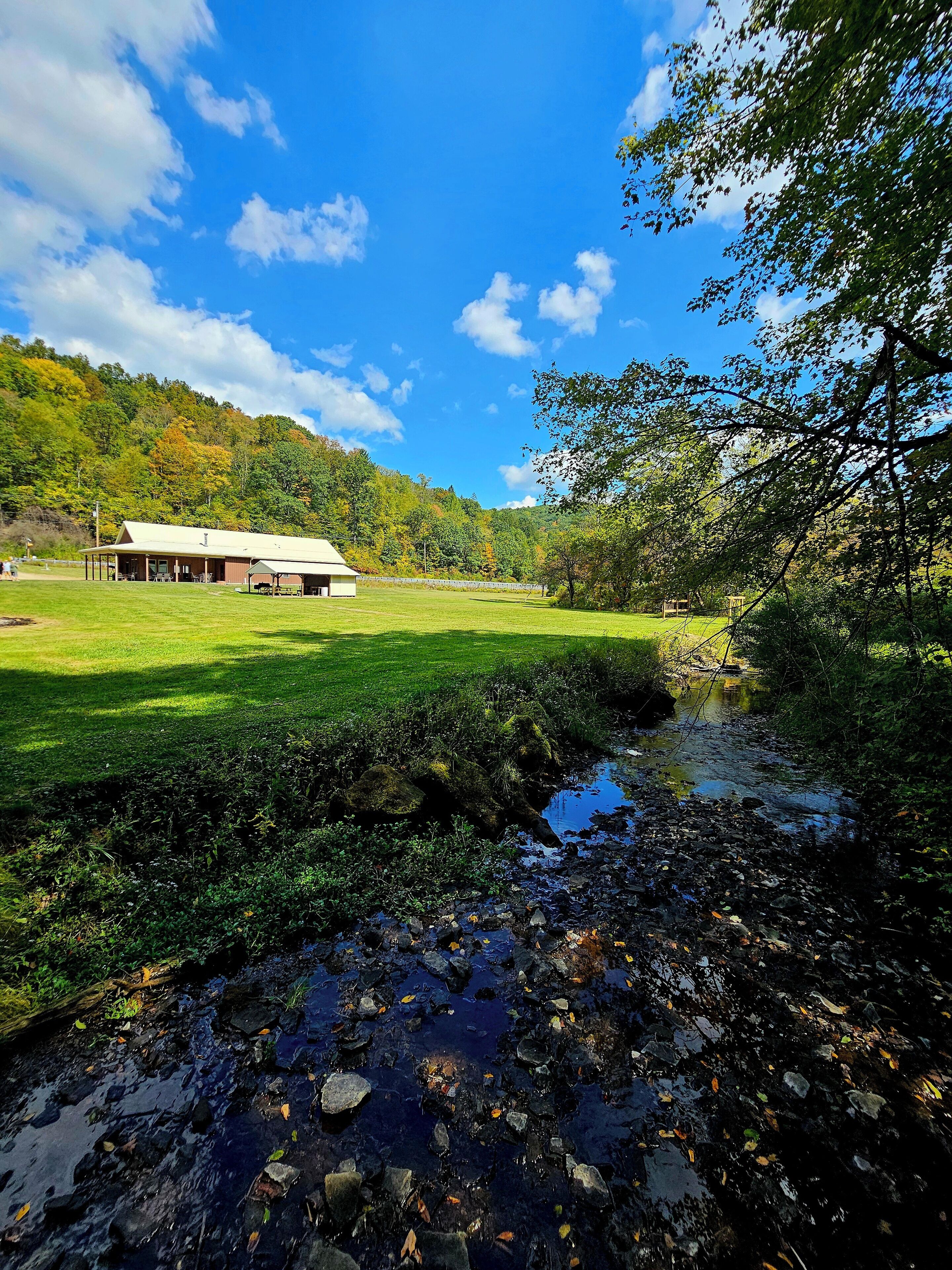 A rustic building with a large porch and a covered area, surrounded by a lush green field and a small stream.