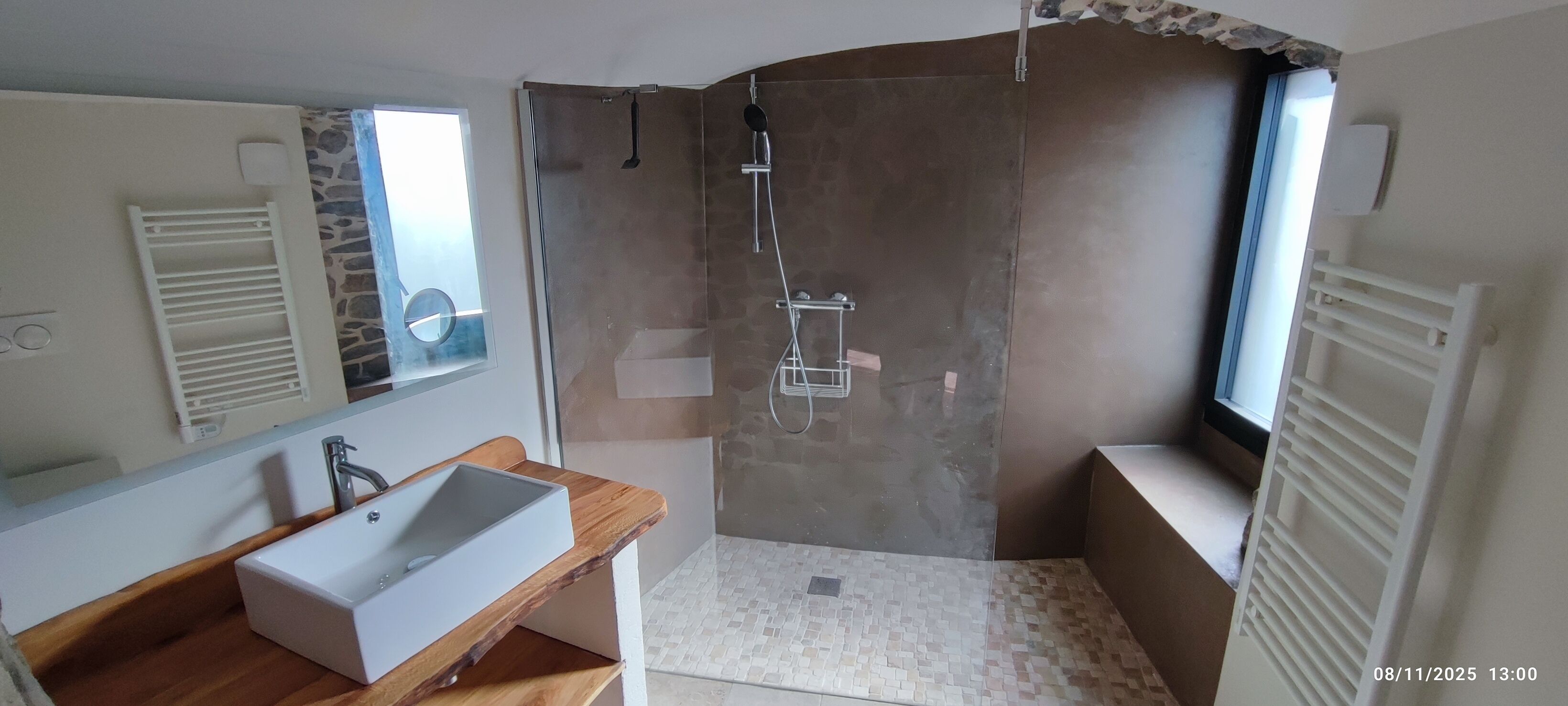 Une salle de bain dotée d’un grand miroir, d’un meuble-lavabo en bois, d’un lavabo blanc, d’une douche avec une porte en verre et d’une fenêtre laissant entrer la lumière naturelle.