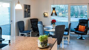 A modern living room with a dining area, featuring a large window with a view of trees and a road, a coffee table with a potted plant, and several chairs. - Awesome home in Ringkøbing with WiFi (Ringkøbing)