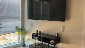 A modern living room with a large flat-screen TV mounted on the wall, a black entertainment unit below, and a window with blinds. - Cozy 1-bedroom apartment in beautiful Vienna with AC and gym access (Vienna)