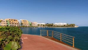 A waterfront promenade with a concrete walkway and wooden railing, leading to a marina with boats. Colorful buildings line the shore, and palm trees are visible in the background. - TAN Residences at Fishing Lodge (Punta Cana)