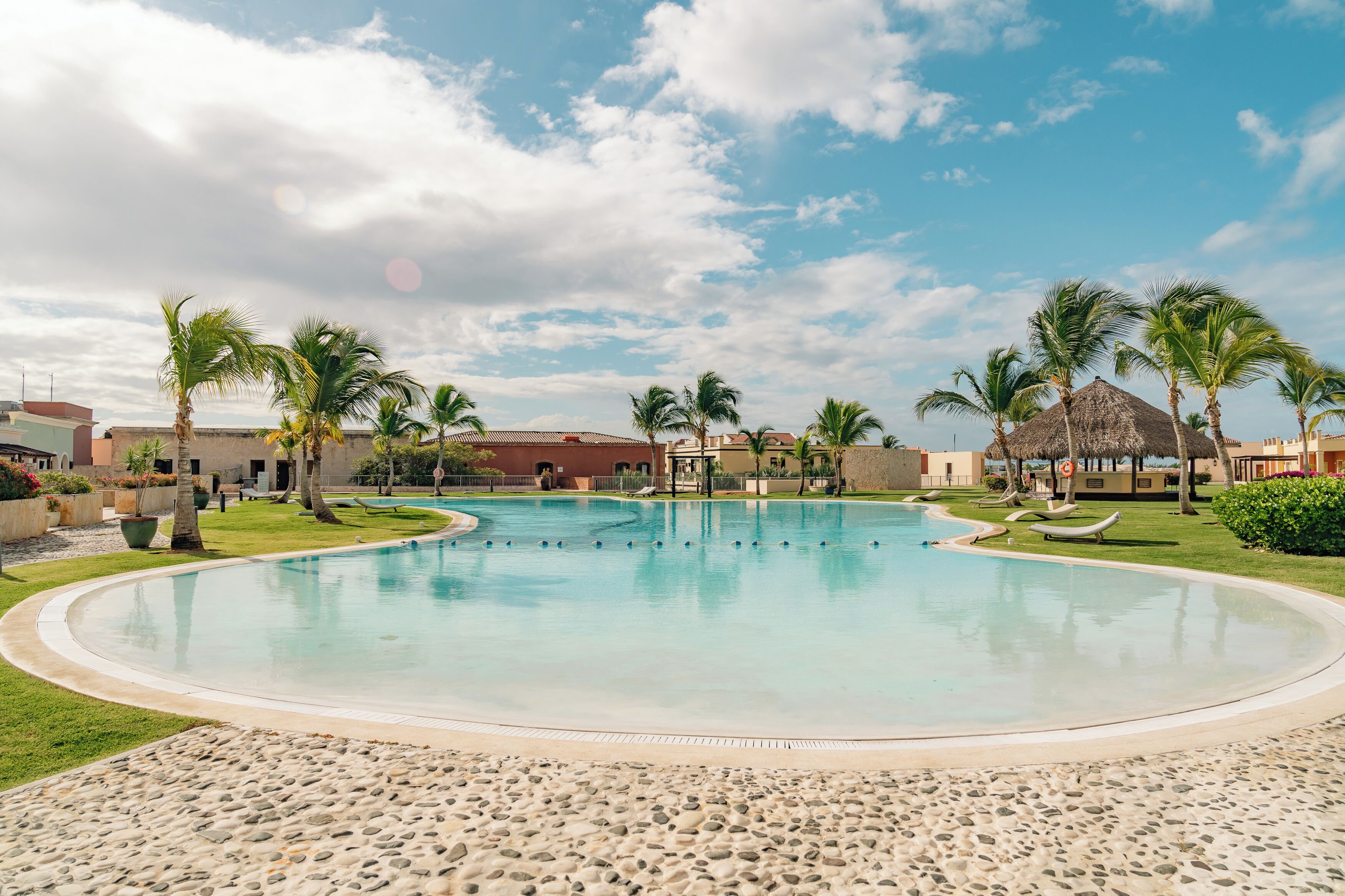 A large, clear swimming pool surrounded by palm trees and lounge chairs, with a building in the background.