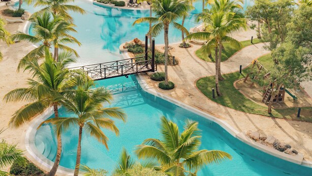 A large, curved swimming pool surrounded by palm trees and a bridge. - TAN Residences at Fishing Lodge (Punta Cana)