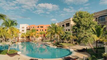 A resort pool area with a clear blue pool, sun loungers, palm trees, and a multi-story building in the background.