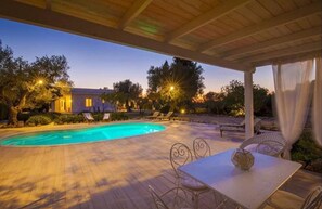 Piscina con sillones de descanso y una mesa bajo una zona cubierta.