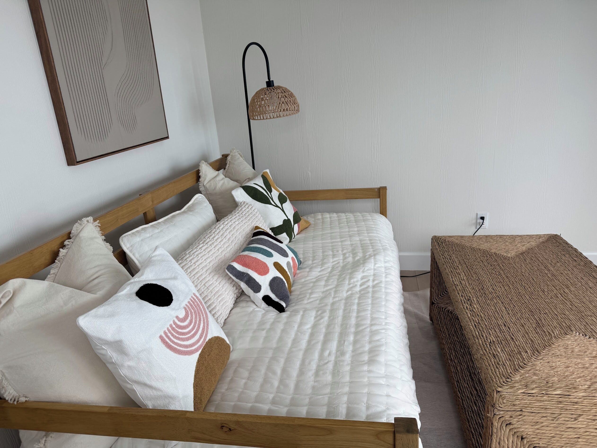 A neatly made bed with a white quilt, multiple pillows, and a woven bench beside it.