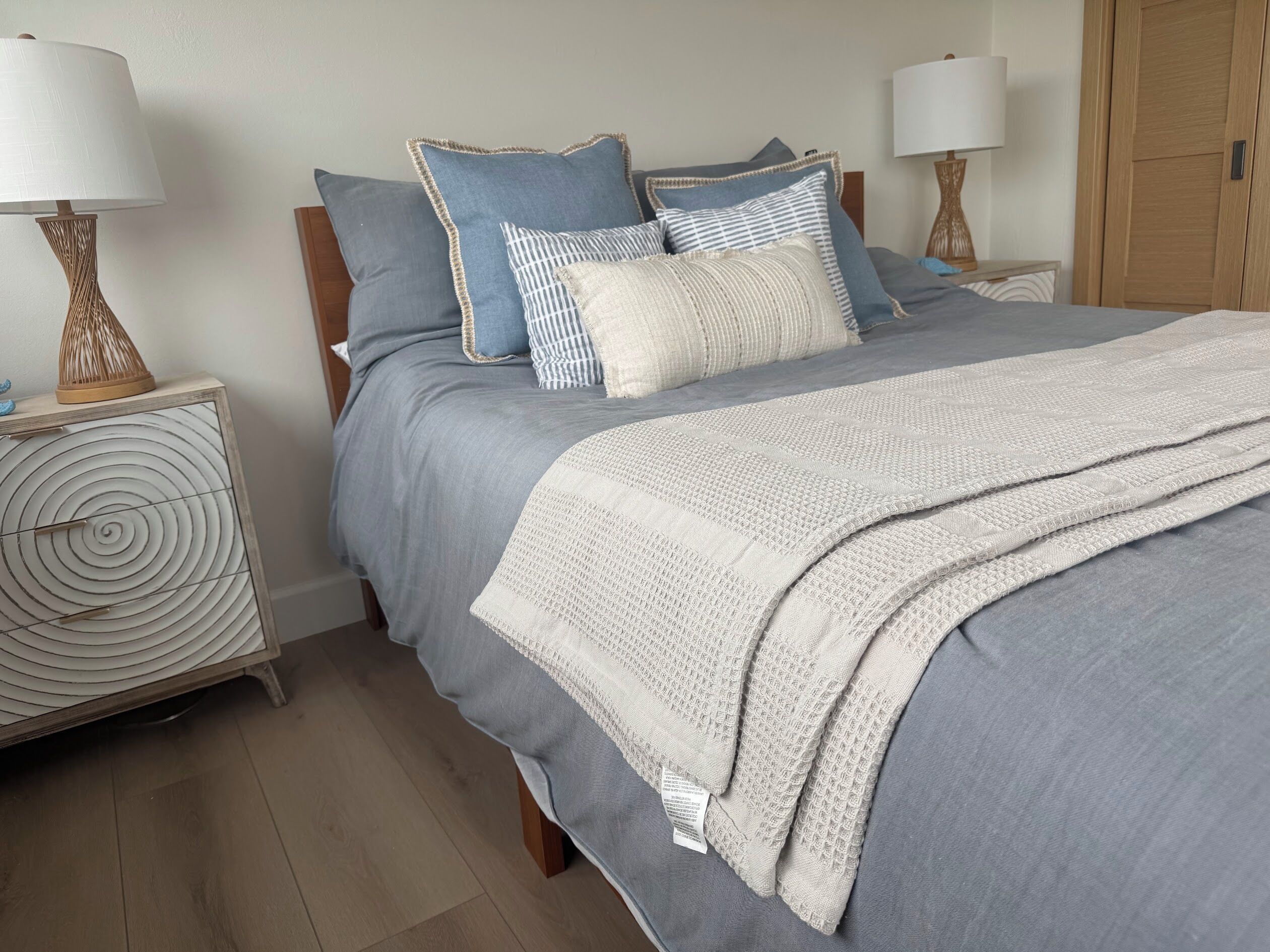 A neatly made bed with a grey duvet and a white textured throw blanket. There are two bedside tables with lamps and a round, spiral-patterned cabinet.