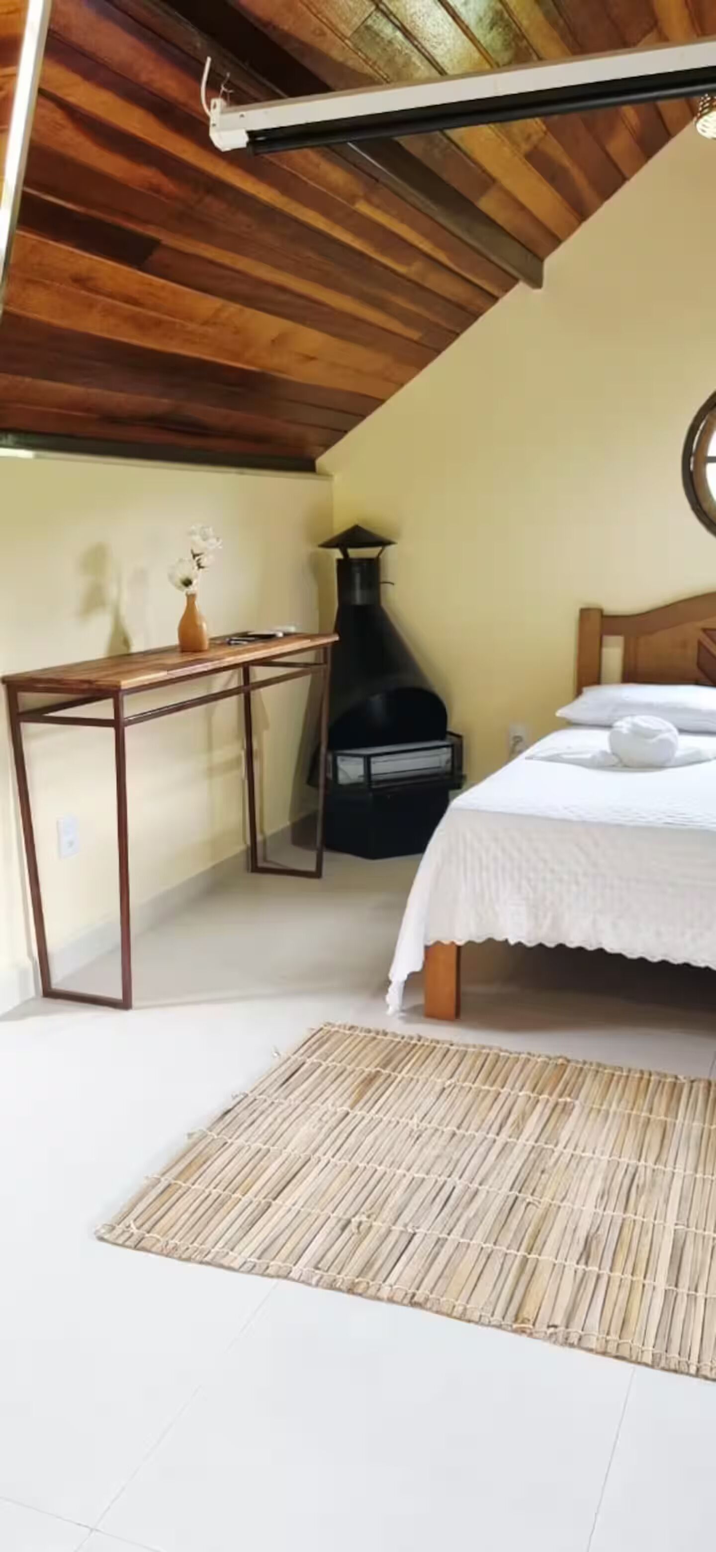 Un dormitorio con cama, una mesa con un florero, un calentador negro y un techo de madera.