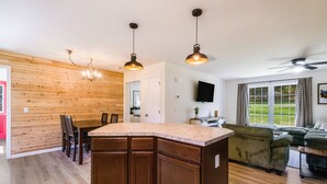 A modern kitchen with wooden cabinetry, a central island, pendant lighting, and a dining area with chairs. - RiverandRidge: 3 Min to River, Hot Tub & Fire Pit (Luray)