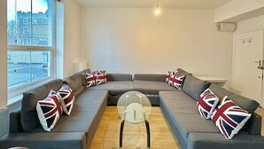 Ein modernes Wohnzimmer mit einem Ecksofa, einem Glastisch und Blick auf eine Stadtlandschaft durch große Fenster.